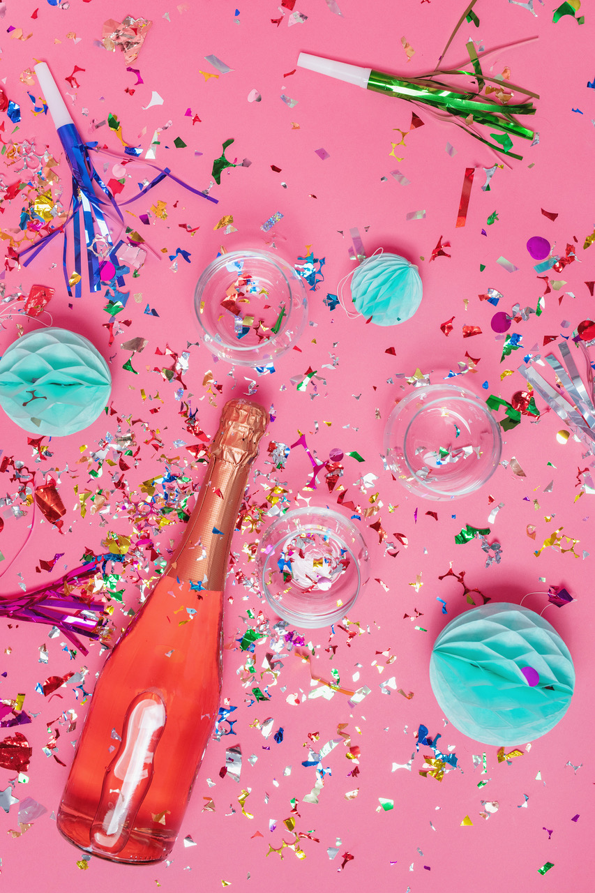 Party equipment; multi-colored confetti,  and a bottle prosecco with a glass on a pink background to symbolize that taking the charge is worth the effort.